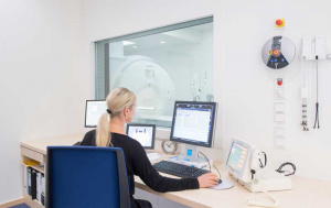 MRT oder CT - junge Frau mit langen blonden Haaren und Pferdeschwanz sitzt mit vor einer Glasscheibe auf einem blauen Bürostühl an einem Schreibtisch. Sie hhat die Hand an einer Computermaus und drei Bildschirme mit MRT Bildern vor sich. Hinter der Glasscheibe sieht man einen hellen Raum mit einem MRT Gerät. Eine weiße große Röhre.