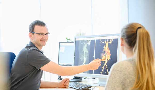 Ein junger Arzt sitzt an einen Schreibtisch. Er trägt ein graues Polohemd und lächelt eine blonde Patientin an, die ihm gegenüber sitzt. Die Patientin sieht man nur von hinten. Sie trägt ein helles Shirt. Er erklärt ihr auf zwei Bildschirmen ein MRT Untersuchungs Bild von einem MRT Blutgefäße und deutet mit einem Kugelschreiber auf ein Blutgefäß am Bildschirm. High-End-Geräte MRT Termin in München auch für Patienten mit Platzangst Die MRT ist ein strahlungsfreies und unschädliches Verfahren zur Schnittbilddiagnostik des gesamten Körpers. Die Untersuchung ist wegen fehlender Strahlenbelastung auch besonders gut für junge Patienten, Kinder und für Schwangere ab dem 3. Schwangerschaftsmonat geeignet. 