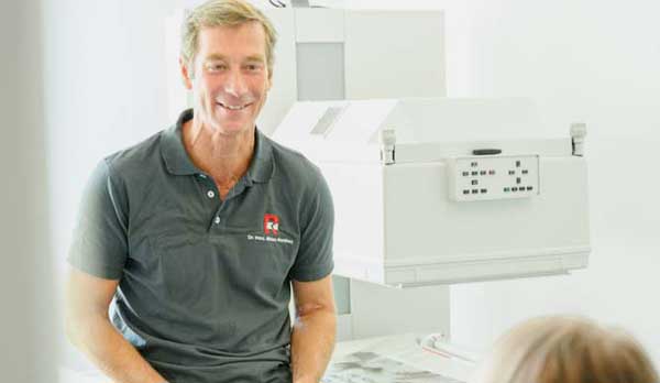 Dr. manthey NUK Myokardszintigraphie. Mittel alter Mann sitzt halb stehend angelehnt auf einer Behandlungsliege mit weißem Papier. Er trägt ein graues Polohemd mit Radiologie Logo und lächelt eine Patientin an, die vor ihm sitzt. Er hat blonde kurze Haare und sonnengebräunte haut.