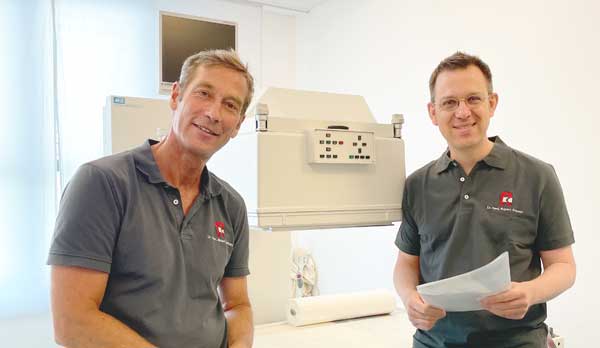 Nierenszintigraphie: Dr. Manthey und Dr. Schmid stehen vor einem weißen Behandlungsgerät, beide tragen graue Poloshirts und besprechen einen Nuklearmedizinischen Befund zur NUK Nierenszintigraphie. Der rechte Mann hält die Unterlagen vor sich in der linken Hand. Der linke Mann hat blonde Haare und sonnengebräunte Haut, der rechte Mann dunkle kurze Haare. Beide blicken freundlich in die Kamera