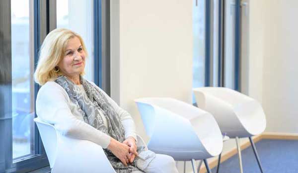 Skelettszintigraphie: eine ältere Frau sitzt in einem Wartezimmer mit blauem Boden auf weißen Stühlen mit Metallbeinen. Im Hintergrund große bodenhohe Fenster mit grauen Fensterrahmen und eine weiße Wand. Die Frau trägt ein helles Shirt und einen grau-blaues Tuch. Sie hat rot-blonde haare und lächelt in den Raum hinein