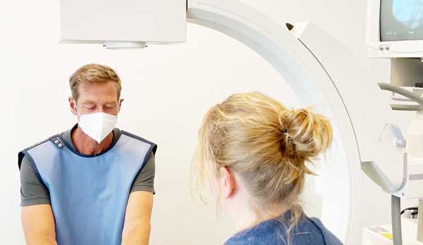 Schmerztherapie. Dr. Manthey München. NUK Untersuchungen und Therapien. Ein blonder mittelalter Mann mit mundschutz und grauem Polohemd trägt eine hellblaue Bleischürze und sitzt lächelnd einer Patientin am NUK Gerät gegenüber. die Patientin hat blonde haare zu einem dutt gebunden und ein dunkelblaues Shirt an