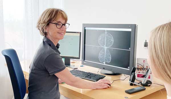 Dr. Birgit Denner: Eine braunhaarige Ärztin sitzt an einem Schreibtisch vor zwei Bildschirmen. Sie dreht sich nach rechts zu einer blonden Frau, die Patientin ist für Brustdiagnostik. Auf den Bildschirmen sieht man eine Mammographie und MRT der Brust. Die beiden sehen sich freundlich an