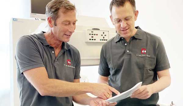 Dr. Manthey und Dr. Schmid stehen vor einem weißen Behandlungsgerät, beide tragen graue Poloshirts und besprechen einen Nuklearmedizinischen Befund zur NUK Lungenszintigraphie. Sie halten die Unterlagen vor sich in beiden Hännden. Der linke Mann zeigt auf etwas auf dem Papier. er linke Mann hat blonde Haare und sonnengebräunte Haut, der rechte Mann dunkle kurze Haare.