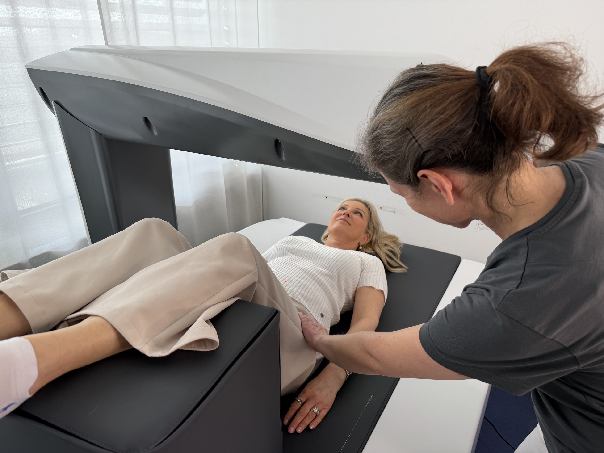 Im Bild sieht man eine Patientin bei der Knochendichtemessung in der Praxis, eine MTA kontrolliert die Einstellungen des Gerätes. Knochendichtemessung DXA München