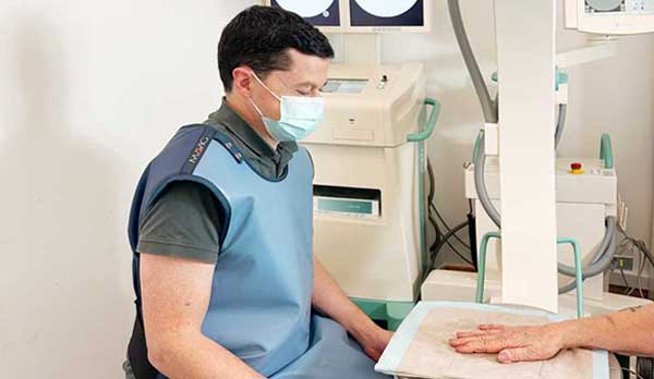 Schmerztherapie. Dr. med. Rupert Schmid in der Radiologie München Süd West. NUK Untersuchungen und Therapien. Ein braunhaariger mittelalter Mann mit Mundschutz und grauem Polohemd trägt eine hellblaue Bleischürze und sitzt lächelnd einer älteren Patientin am NUK Gerät gegenüber, von der nur die rechte Hand zu sehen ist.